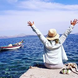 Rear view of woman with arms raised sitting on pier against sky