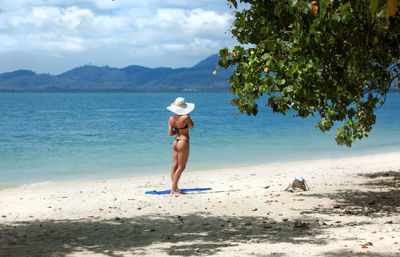 Rear view of sexy slim woman standing at tropical beach against sky and sea