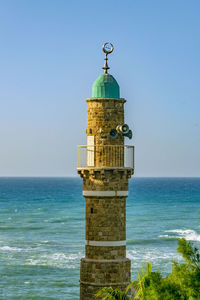 Lighthouse by sea against clear blue sky