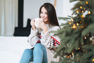 Young asian woman in sweater  with cup of cocoa with marshmallow in room with christmas tree at home