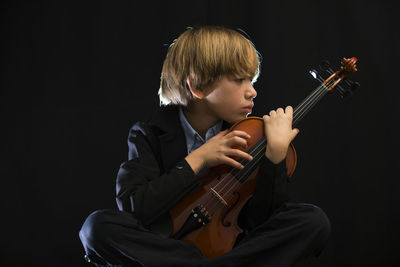 Boy holding violin while sitting against black background