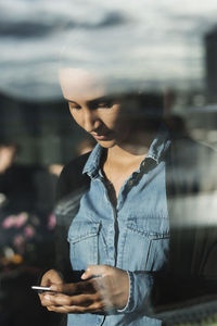 Close-up of young woman using smart phone seen through glass