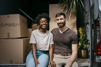 Portrait of happy woman sitting with boyfriend in van trunk full of boxes