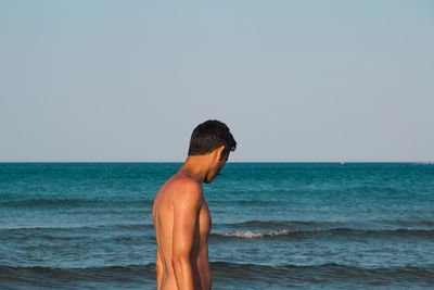 Side view of shirtless teenage boy standing at beach against clear sky