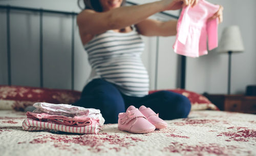 Pregnant woman holding baby clothing while kneeling on bed at home