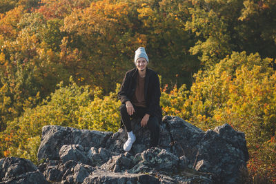 Attractive young man with smile on his mouth in a black coat and grey cap walks sits on the rock