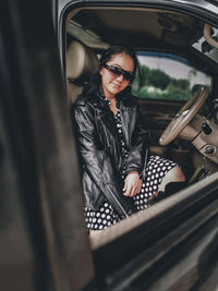 A girl wearing a polkadot dress, jacket, sun glasses and high boots sitting in a car 