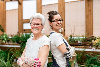 Side view of positive elderly and adult women in gloves and glasses smiling for camera and crossing arms while working in indoor garden together