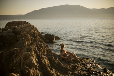 Rear view of man sitting on rock at beach against sky during sunset