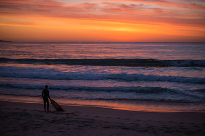 Silhouette surfer with skateboard at beach against sky during sunset