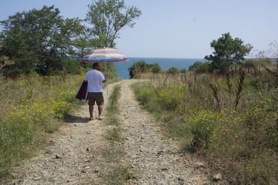 Rear view of man with parasol standing on field by lake during sunny day