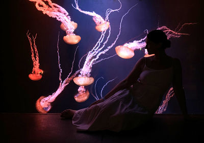 A woman sitting in front of dark aquarium with colored jellyfishes