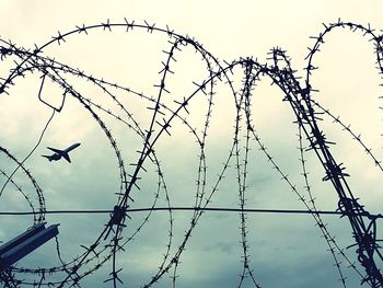 Close-up of barbed wire fence against airplane flying in sky