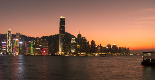 Illuminated buildings by river against sky during sunset