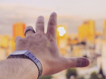 Cropped hand of man gesturing against city during sunset