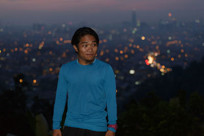 Man looking away while standing against illuminated city at dusk