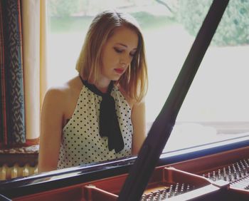 Young woman playing piano against window on sunny day