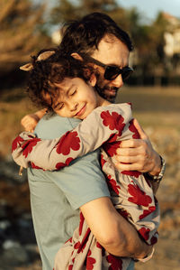 Side view of adult man in sunglasses carrying and embracing girl with closed eyes on weekend day in countryside