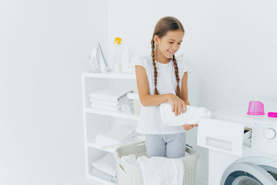 Cute girl holding detergent standing by washing machine at home