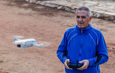 Side view of old man flying the drone looking at the drone while standing against the trees in the park