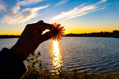 Close-up of hand against orange sky over lake during sunset