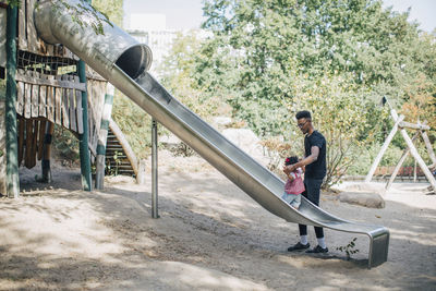 Father holding hands of daughter walking on slide at playground