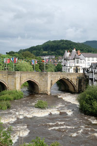 Bridge over flowing river by houses against sky