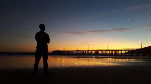 Silhouette man standing at beach against pier during sunset
