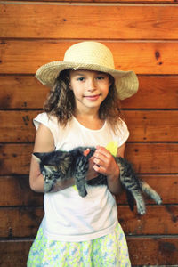 Portrait of cute little girl wearing straw hat holding kitten standing against wooden planks in barn