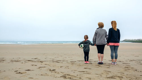 Rear view of woman standing with mother and daughter at beach against sky