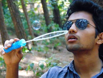 Close-up of young man wearing sunglasses blowing bubbles in forest