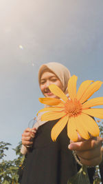 Young woman holding yellow flower against sky