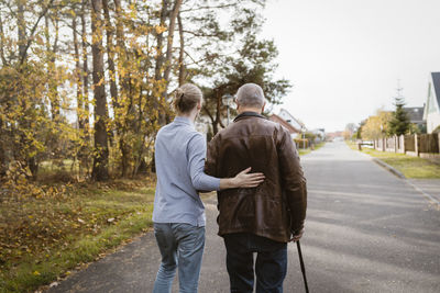 Rear view of caretaker supporting retired senior man walking with cane on road