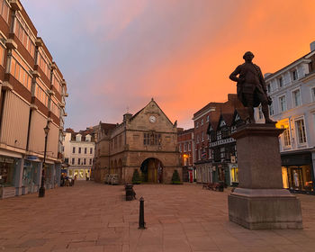 Statue on street amidst buildings against sky at sunset