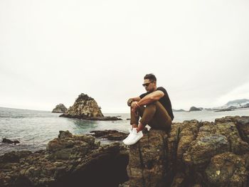 Man sitting on rock by sea against sky