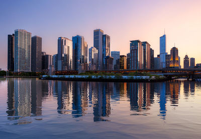 Illuminated buildings by lake in city against sky at dusk