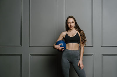Portrait of young woman with medicine ball standing against wall