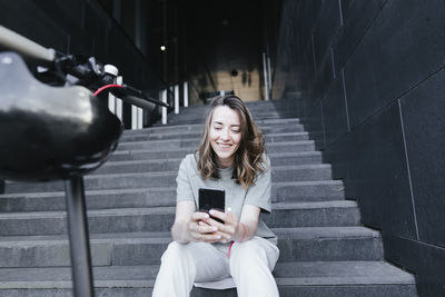 Woman with e-scooter and helmet holding smartphone, sitting on steps