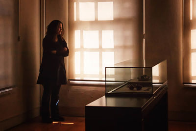 Full length of thoughtful woman standing in living room by sunlight on wall