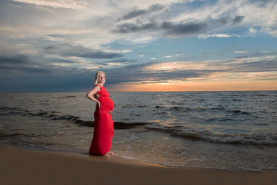 Portrait of pregnant woman standing on shore at beach during sunset