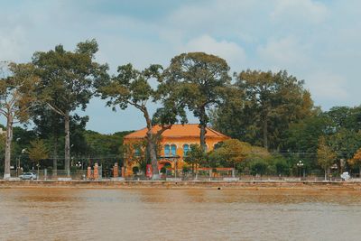 Trees and buildings by river against sky