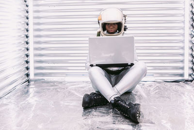 Full body of concentrated african american female in spacesuit typing on netbook while sitting in modern spaceship on light background