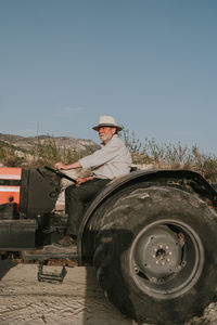 Side view of senior male farmer sitting in tractor while working in agricultural plantation with blooming apricot trees in countryside on summer day looking away
