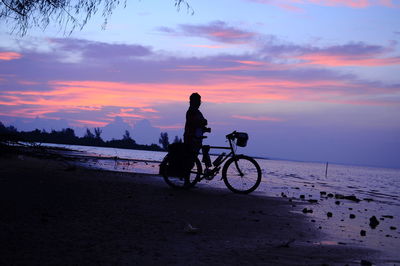 Silhouette man riding bicycle on beach against sky during sunset