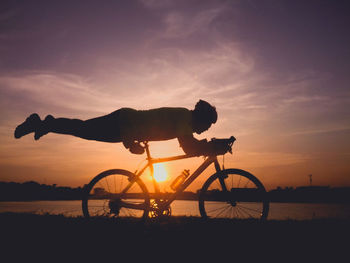 Silhouette man balancing on bicycle seat by lake against sky during sunset