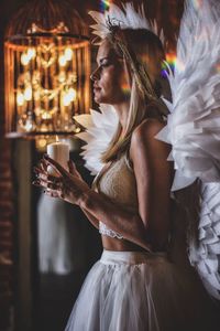 Young woman wearing angel costume holding candle while standing at home