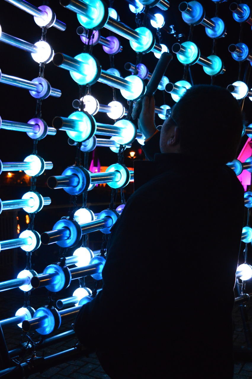 REAR VIEW OF MAN STANDING IN ILLUMINATED LIGHTS