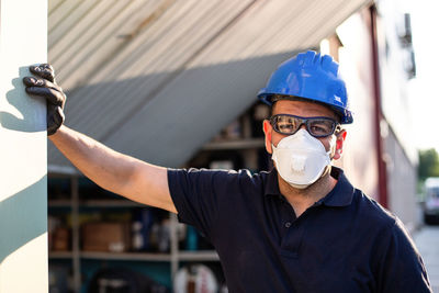 Adult male mechanic in protective glasses and respirator with blue hardhat looking at camera while standing near workshop building on street