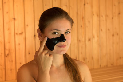 Portrait of beautiful young woman applying facial mask on face while sitting in sauna