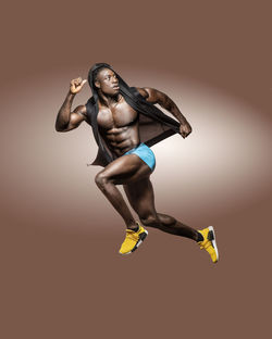 Full length of shirtless athlete jumping against brown background